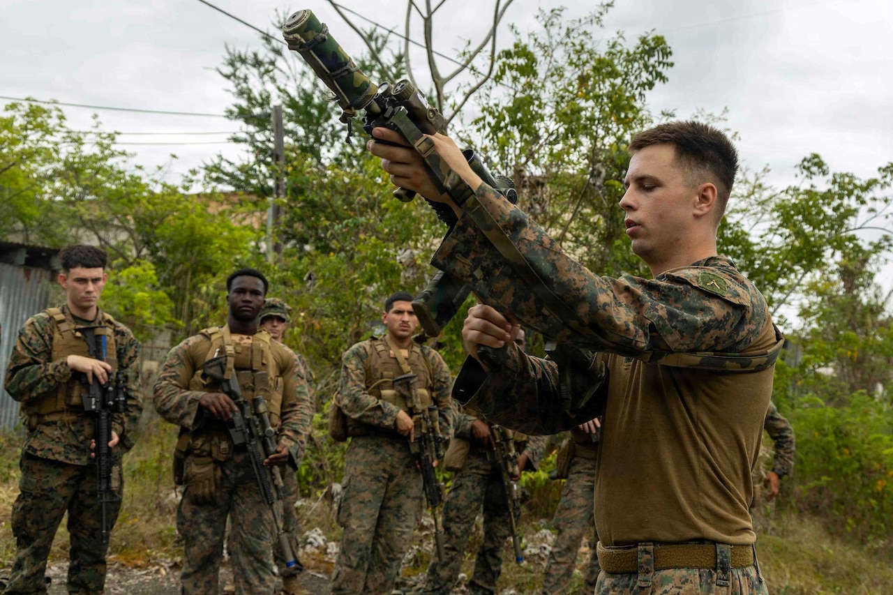 A man wearing a camouflage military uniform holds a rifle in the air while standing outside in a wooded area as several other people in similar attire observe.