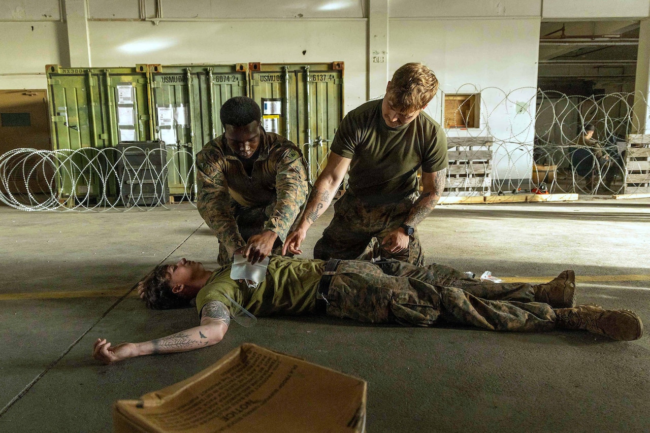 A man wearing a camouflage military uniform checks on a simulated patient lying on the ground as another man in similar attire advises him.