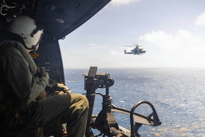 A person wearing a flight suit looks out the open door of a military helicopter at another helicopter flying over the ocean.