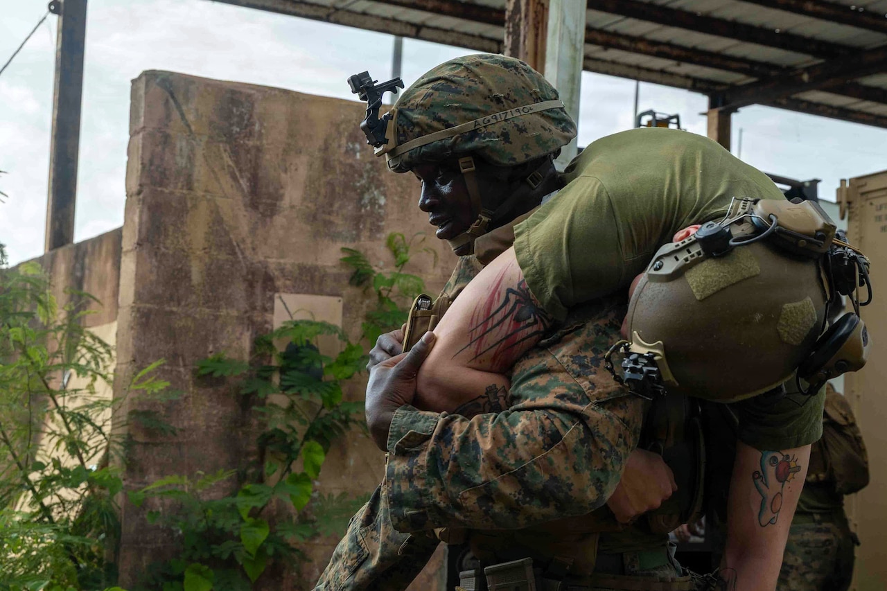 A man wearing a camouflage military uniform carries another man in similar attire over his shoulder while walking outside.
