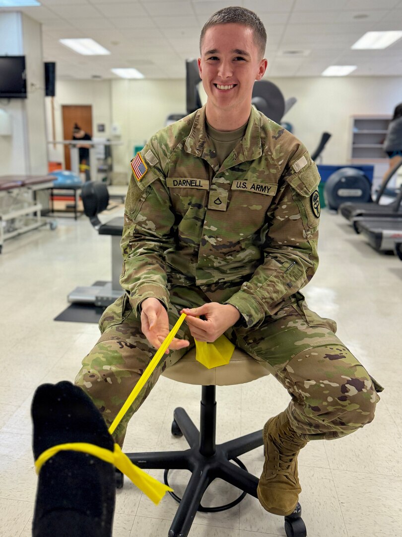 Soldier sitting in front of a patient, holding an yellow elastic band attached to the patient's leg
