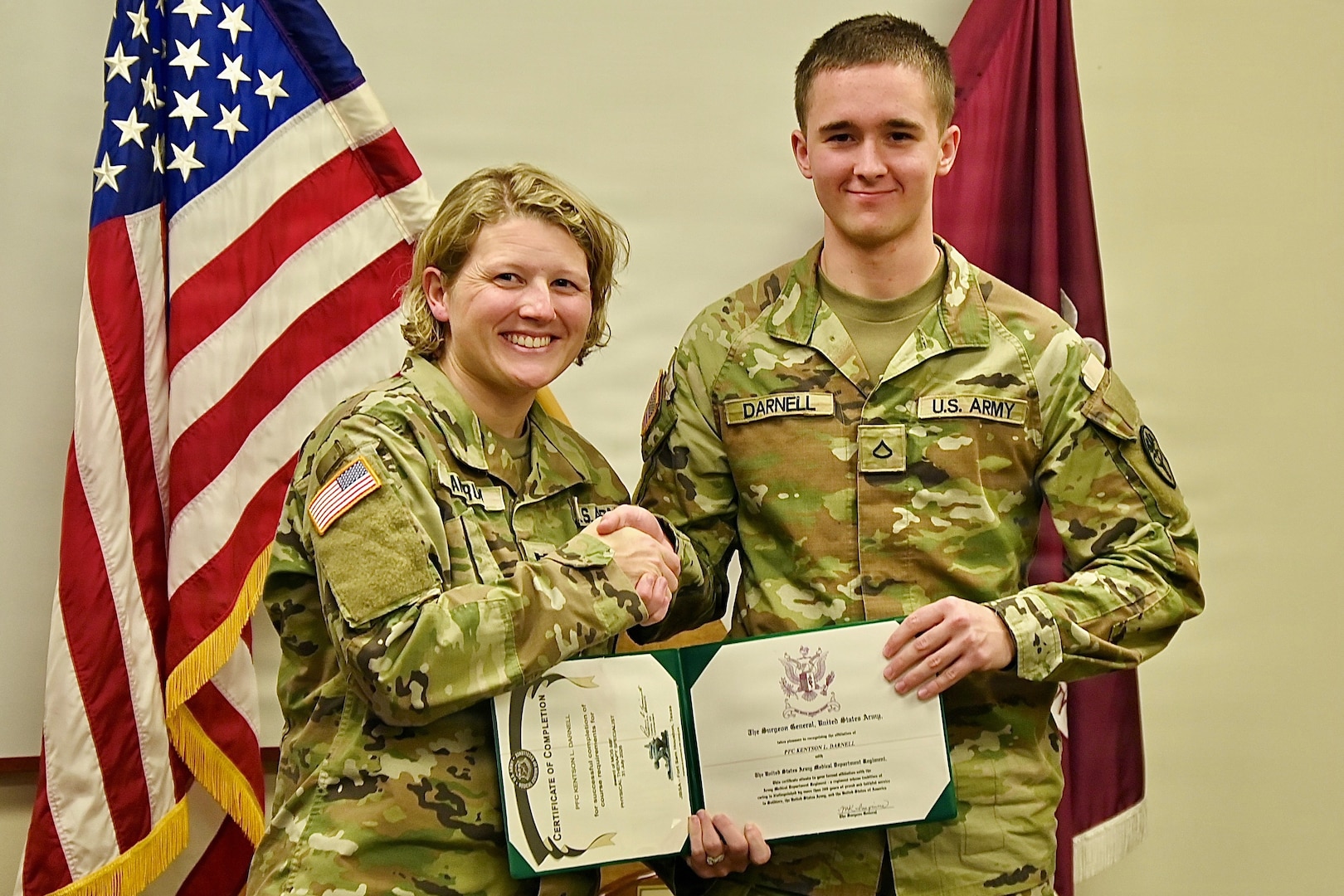Military officer standing in front of flags shaking hands with and presenting a certificate to an enlisted Soldier.