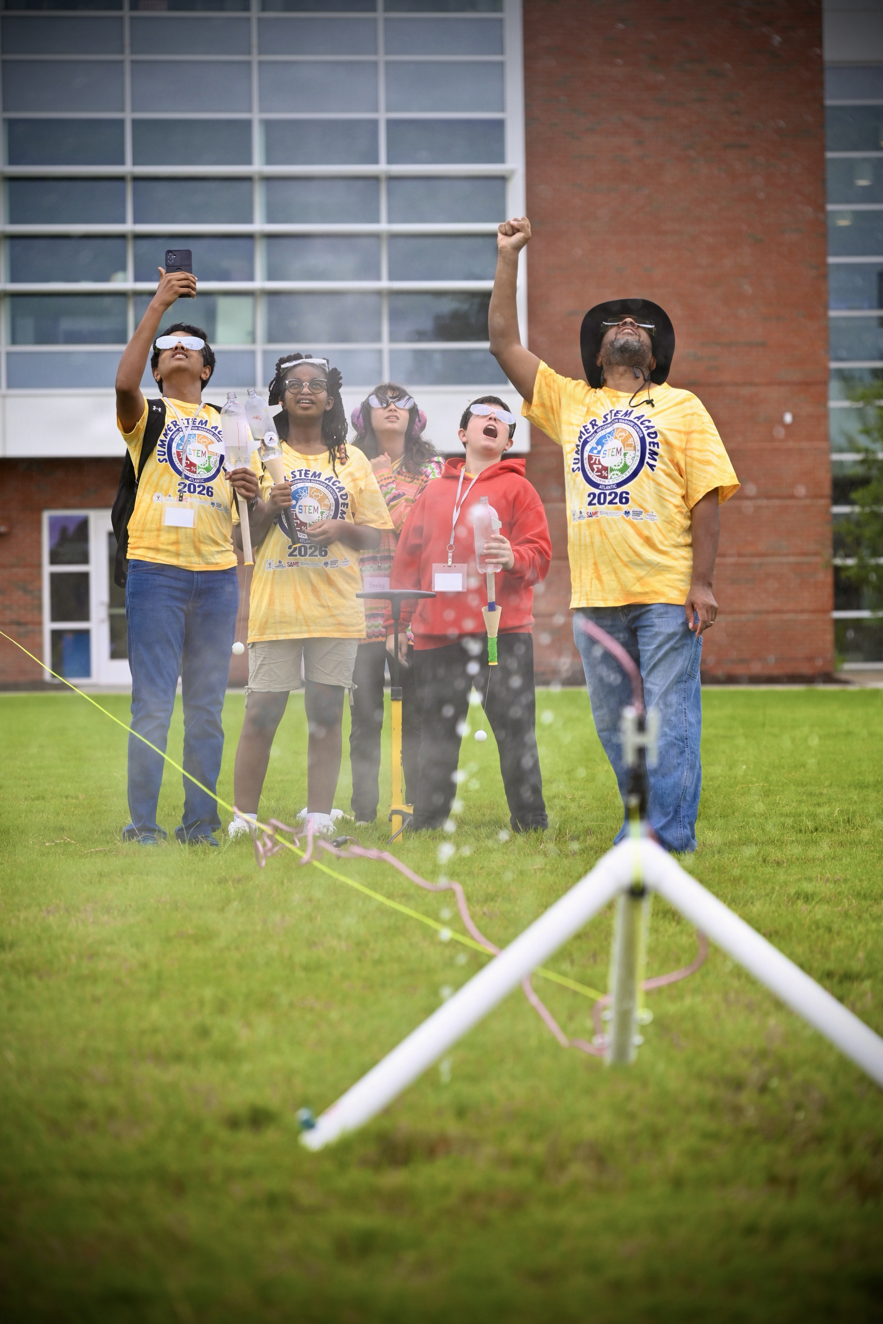 A group of middle school students gathers outdoors on a grassy campus field to watch a water rocket being prepared on a launcher stand during NIWC Atlantic's summer STEM camp.