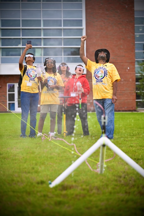 A group of middle school students gathers outdoors on a grassy campus field to watch a water rocket being prepared on a launcher stand during NIWC Atlantic's summer STEM camp.