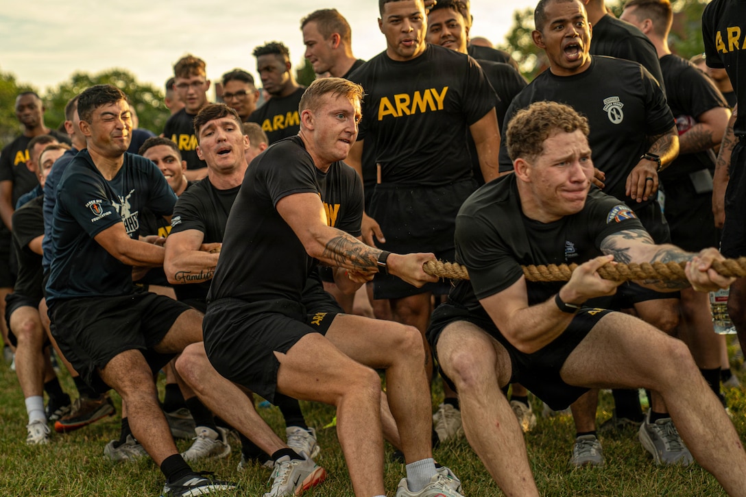 Soldiers in black athletic gear strain while pulling a rope on a field as fellow soldiers watch.