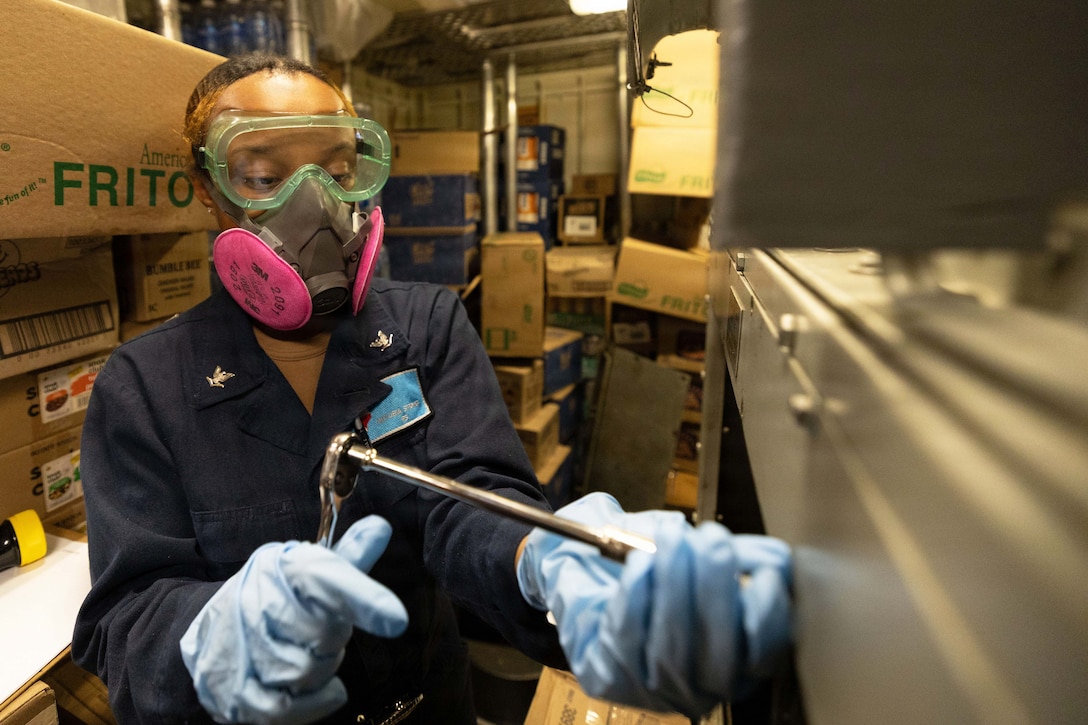 A sailor wearing goggles and gloves uses a tool on a fan coil unit in a storage room stacked with boxes.