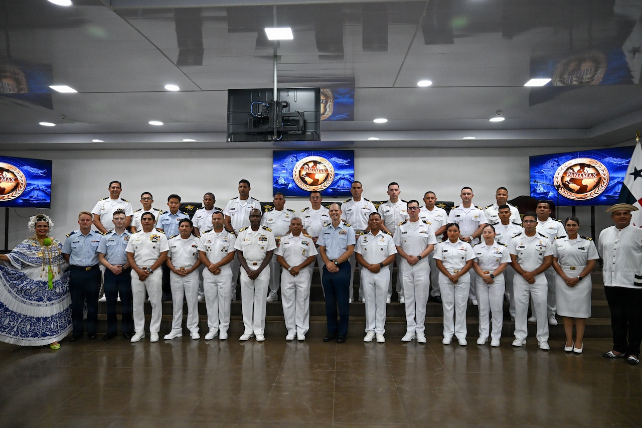 More than a dozen people in various formal military uniforms pose for a group photo in a conference room with monitors on the wall behind them.