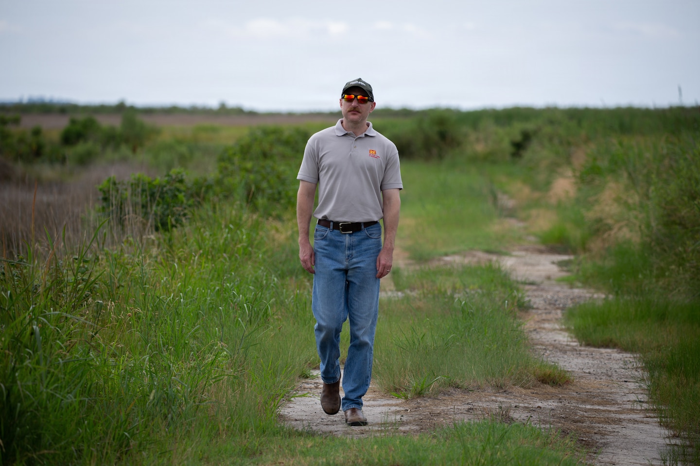 A man wearing a grey polo shirt, blue jeans, a baseball cap, and sunglasses walks along a dirt path in a grassy marsh area.