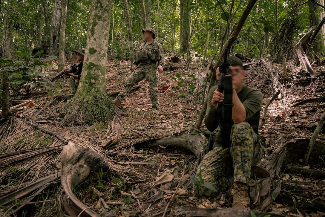 Three men in camouflage military uniforms and holding weapons stand and crouch in a wooded area facing opposite directions.