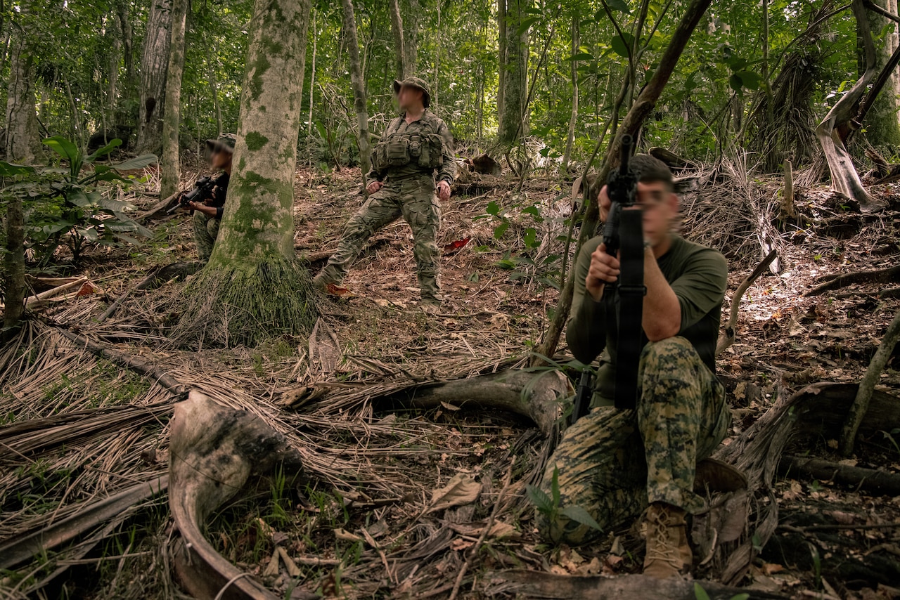 Three men in camouflage military uniforms and holding weapons stand and crouch in a wooded area facing opposite directions.