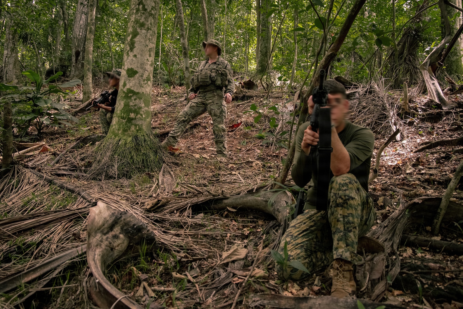 Three men in camouflage military uniforms and holding weapons stand and crouch in a wooded area facing opposite directions.
