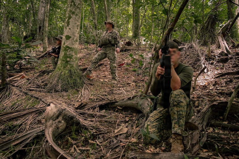 Three men in camouflage military uniforms and holding weapons stand and crouch in a wooded area facing opposite directions.