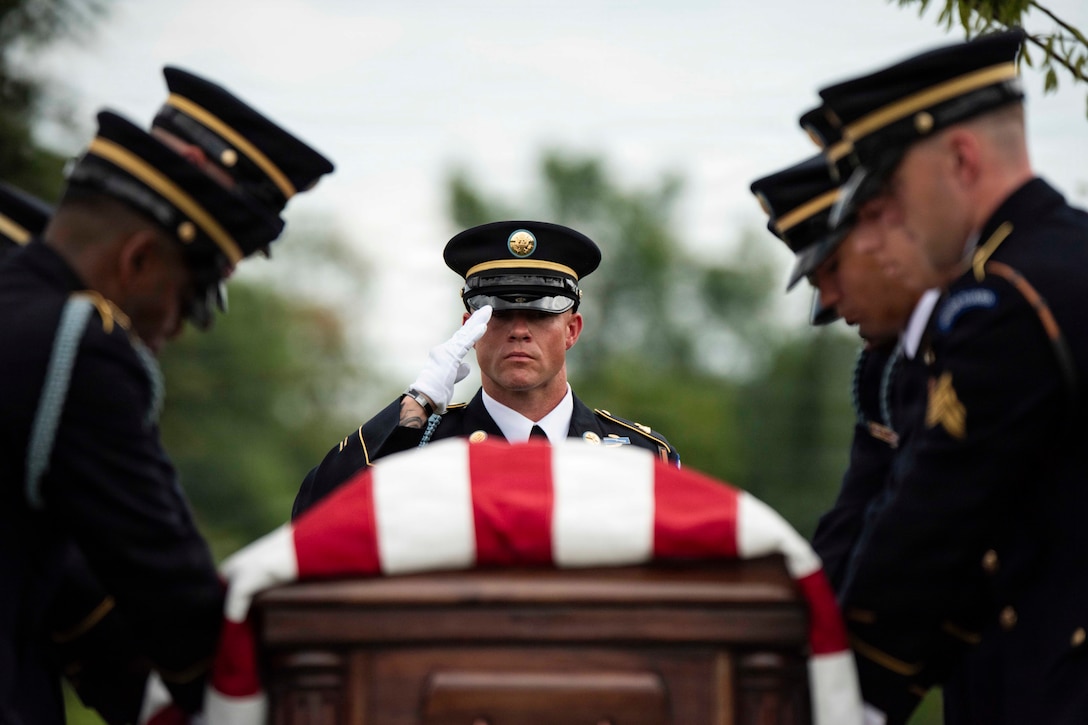 Soldiers in dress uniforms surround a coffin with an American flag draped over it. One soldier standing at the top of the coffin salutes. Trees are in the background.