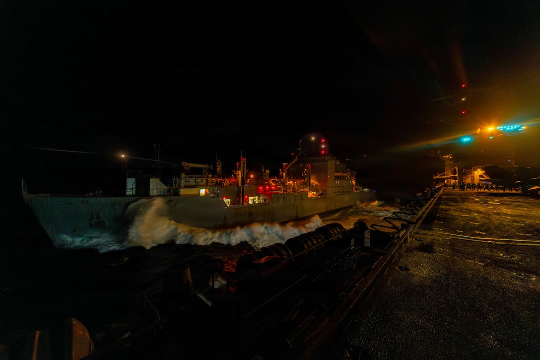 A ship with red and white lights on its deck pulls alongside another ship at night. Water splashes along its side.