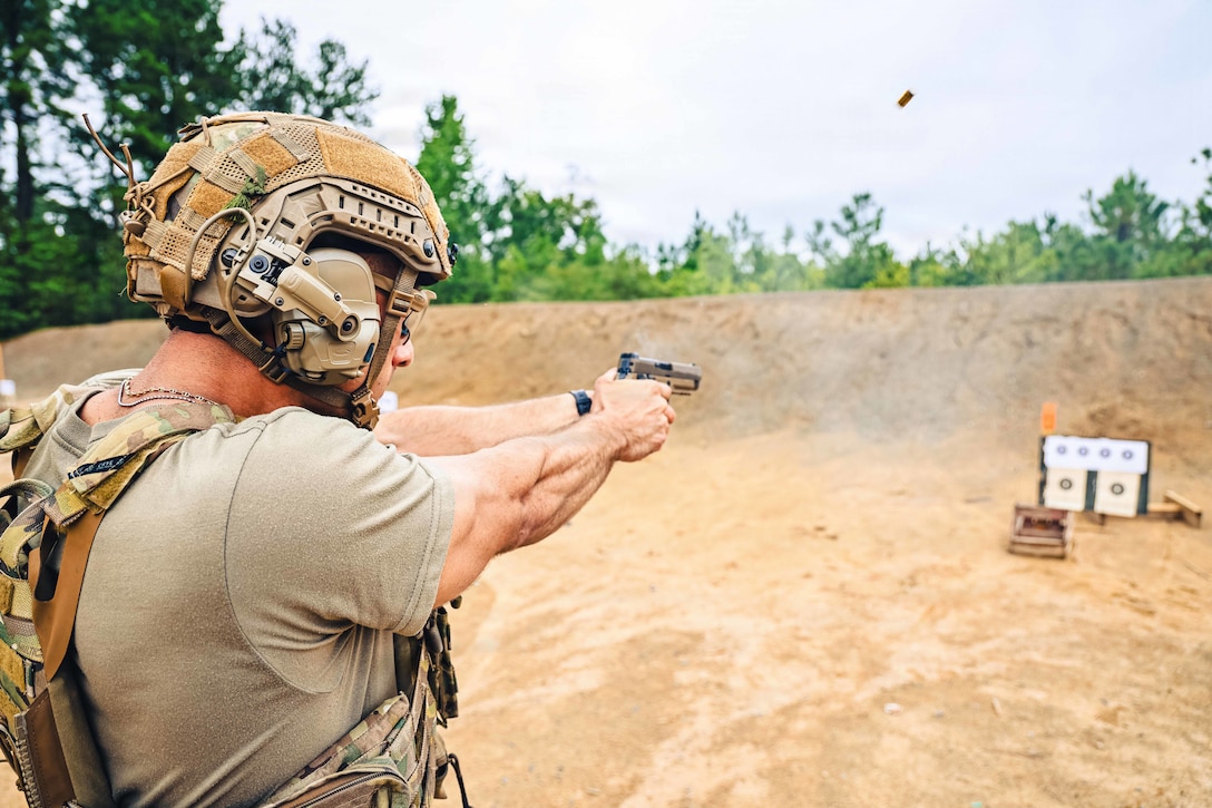 A person in a khaki T-shirt and helmet and a camouflage vest fires a pistol at a target outdoors. A shell casing is in the air.