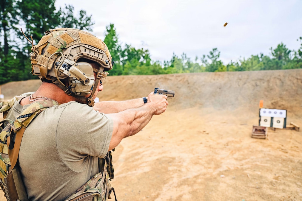 A person in a khaki T-shirt and helmet and a camouflage vest fires a pistol at a target outdoors. A shell casing is in the air.