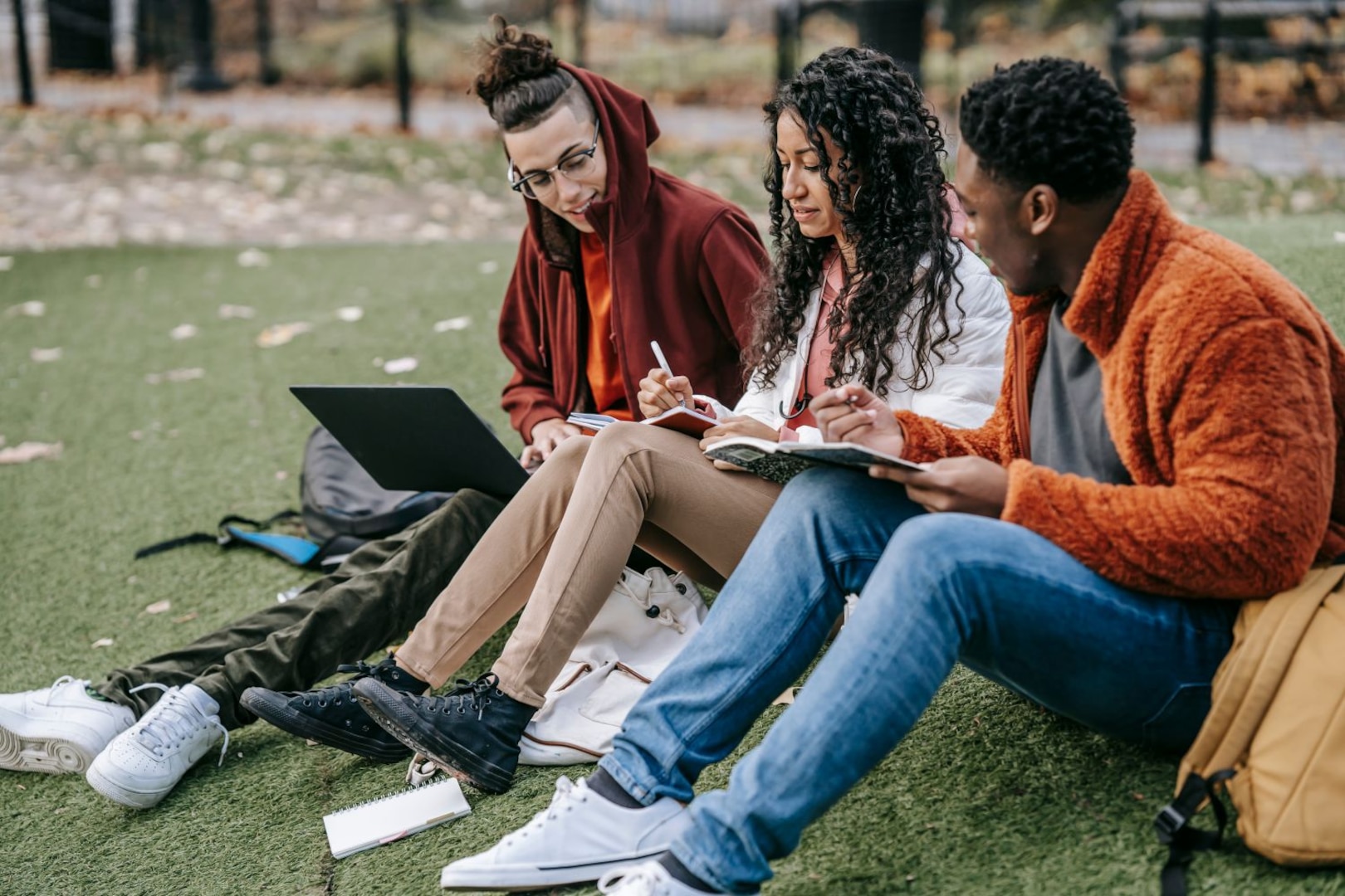 Three people sit on grass while writing on notebooks and a laptop.