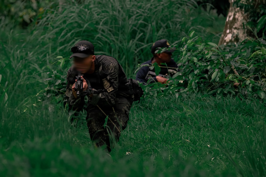 Two people in camouflage military uniforms and holding weapons crouch in a field with tall grass.