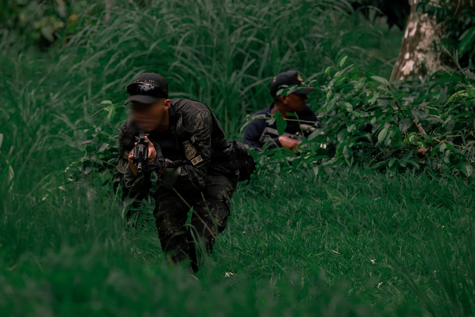 Two people in camouflage military uniforms and holding weapons crouch in a field with tall grass.