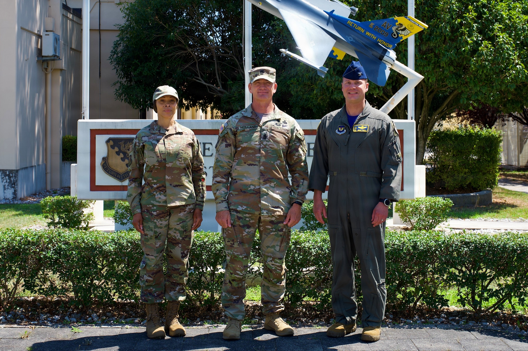 Three service members pose for a photo in front of a the base headquarters.