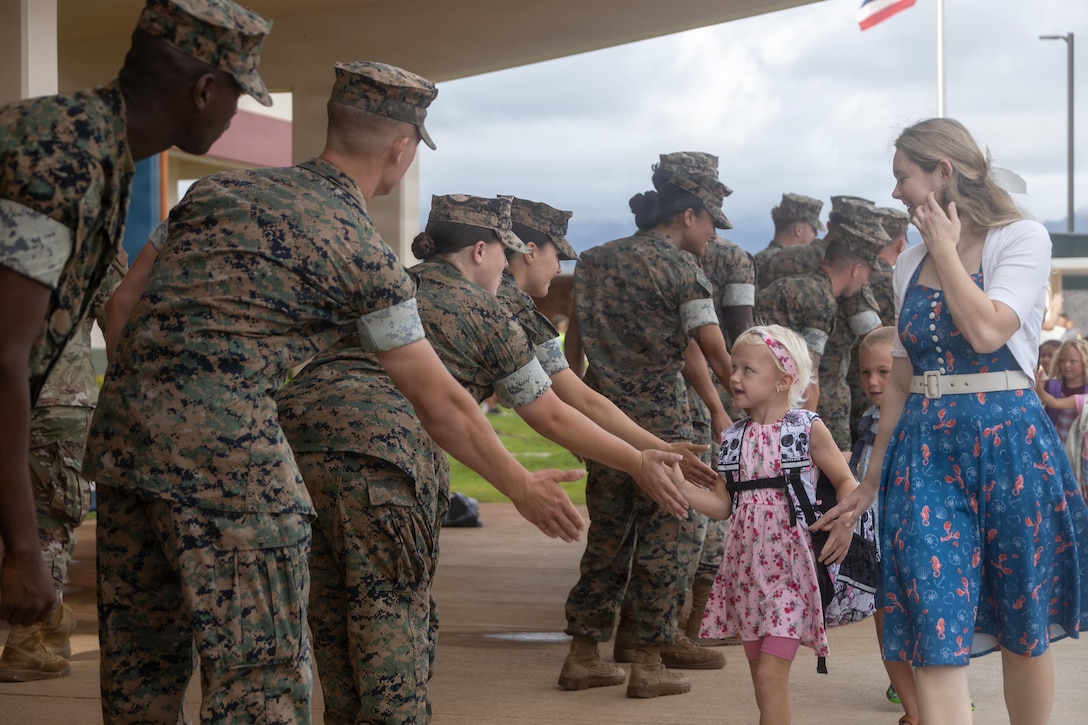 A civilian guides a child as they high-five Marines in camouflage uniforms standing in a line outside of a school.