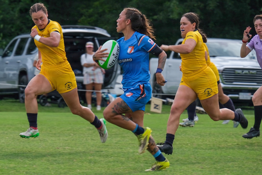 A Coast Guard rugby player in a blue uniform runs with the ball on a field while being chased by Navy players in gold uniforms as a spectator watches from the trunk of their car in the background.