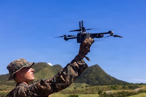 A man wearing a camouflage military uniform holds a military drone in the air as he stands in a grassy field with rolling hills in the background.