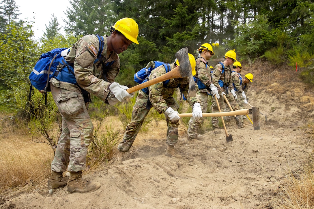 Six people in camouflage military uniforms and hard hats use tools to dig a clearing in the ground.