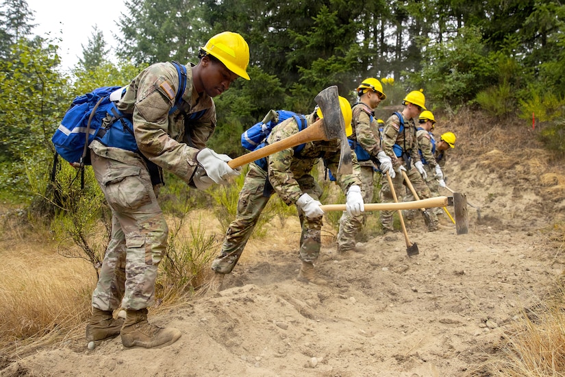 Six people in camouflage military uniforms and hard hats use tools to dig a clearing in the ground.