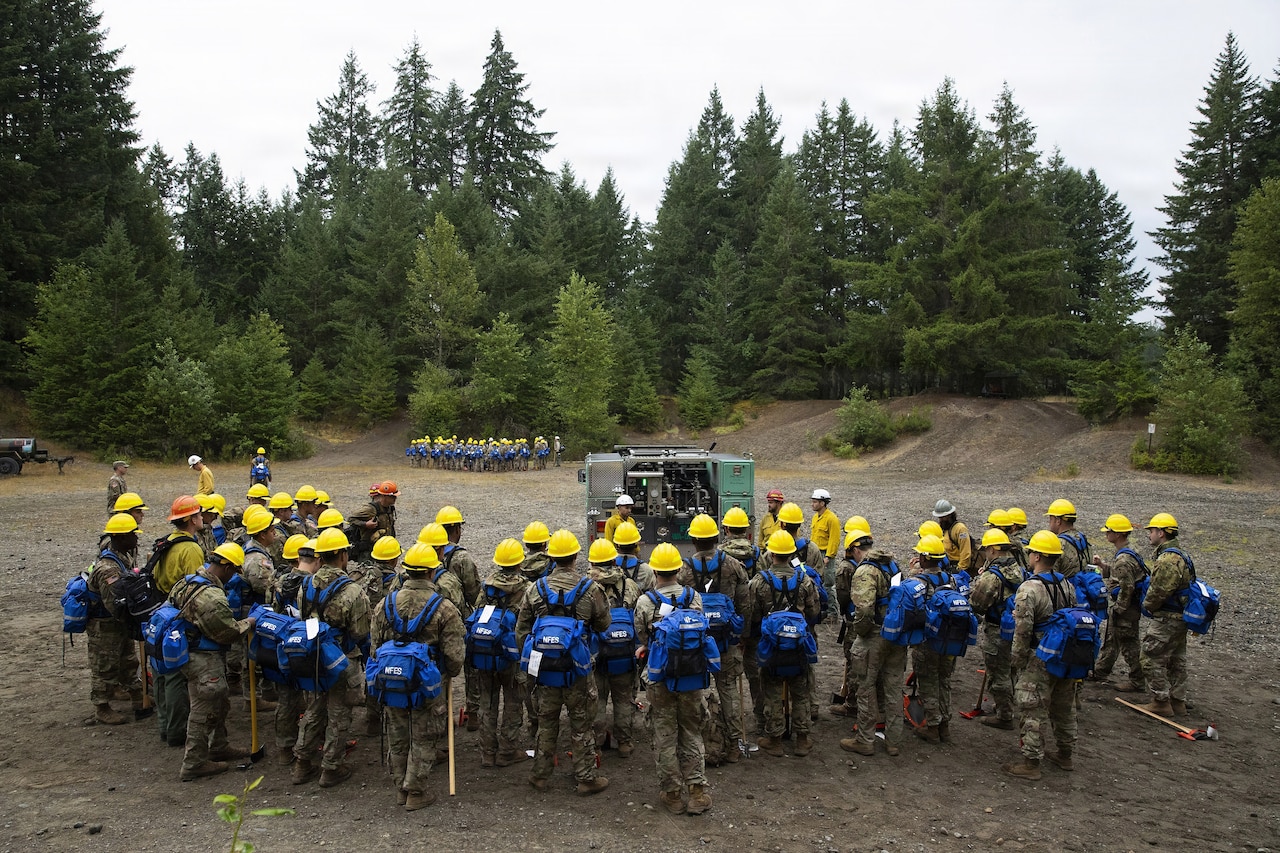 Dozens of people in camouflage military uniforms and hard hats stand in a clearing in the woods as they listen to another person in a hard hat speak.