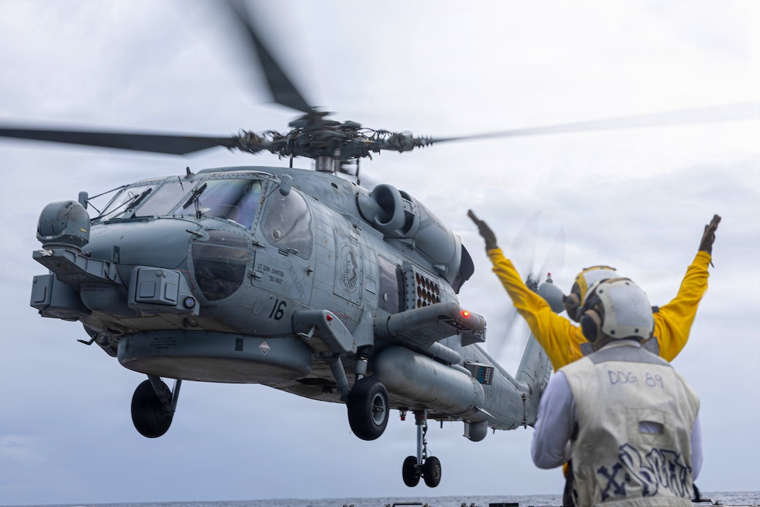 A person in a yellow shirt and white and yellow helmet raises their arms in a V-shape while looking up at a gray military helicopter. Water is in the background and another person is behind the signaler.