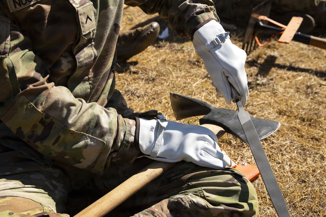 A close-up of a person in a camouflage miliary uniform sitting on the ground sharpening an axe.