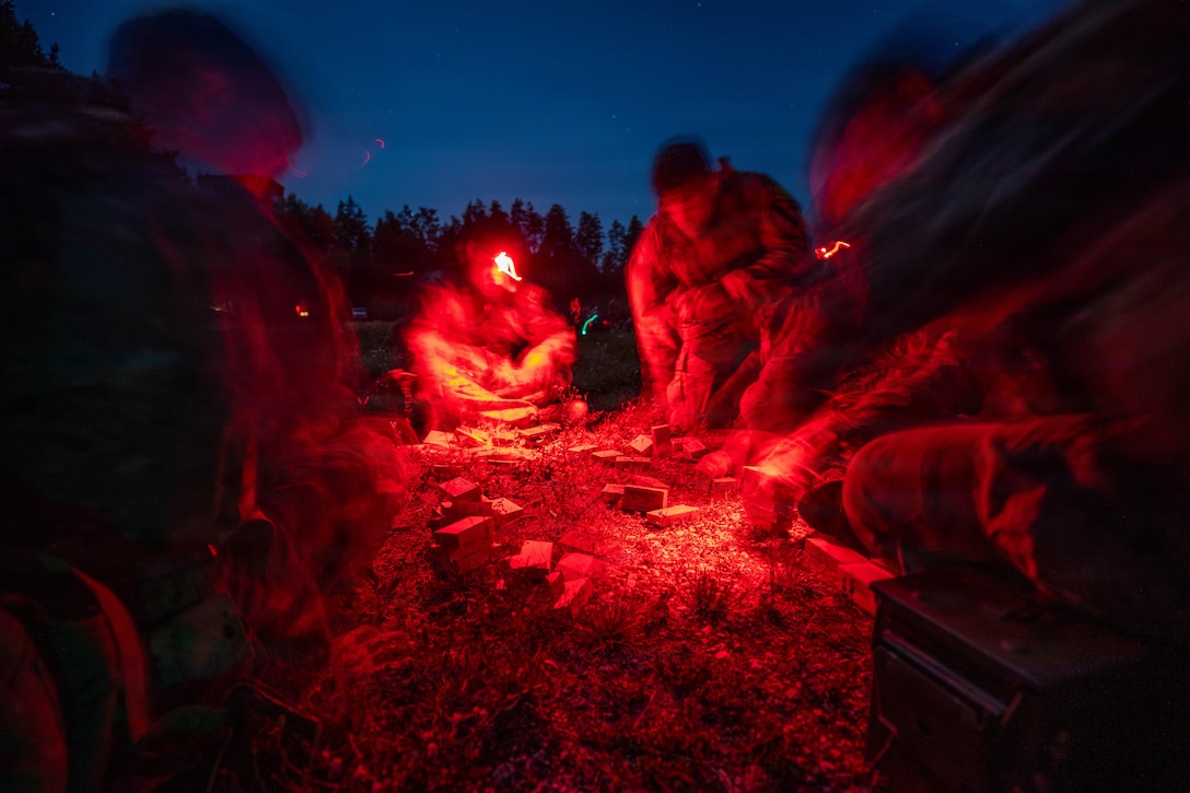 Soldiers in a circle on the ground use red-lens headlamps and separate ammunition. Trees are in the background.