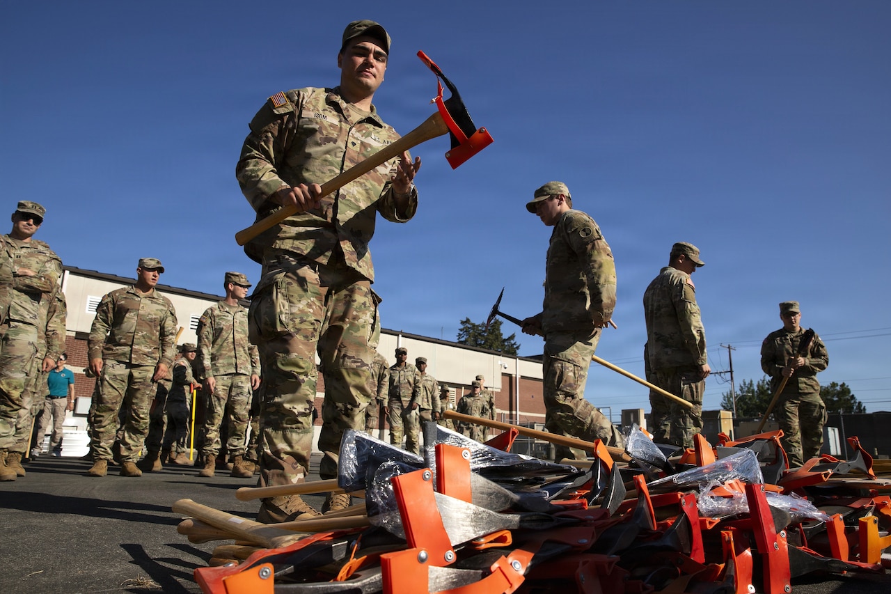 A man in a camouflage military uniform holds an axe in his hands as several other people in similar attire wait behind him to pick up an axe off the ground.