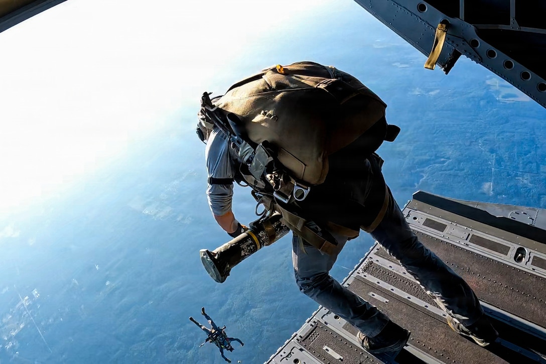 A person wearing a backpack leans over a helicopter's open hatch. Another person is in a free fall to the ground below.