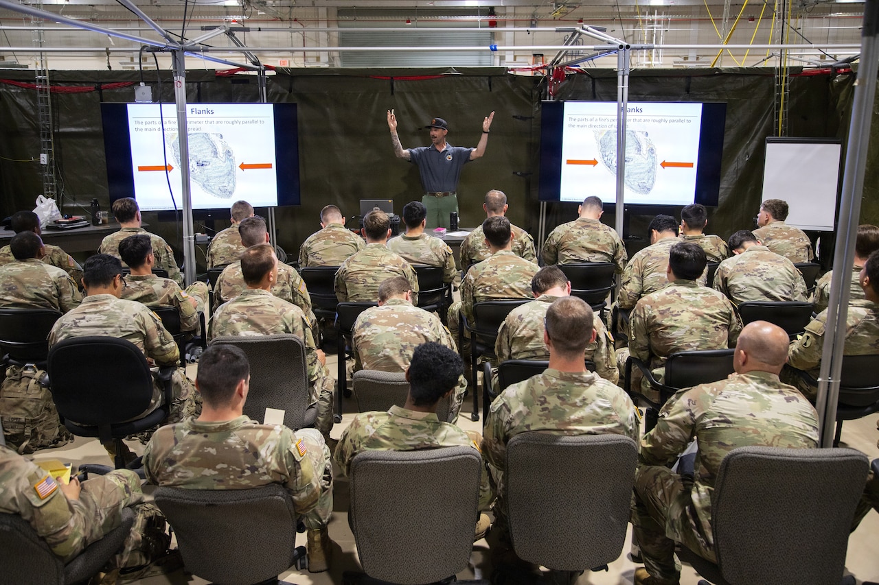 Dozens of people in camouflage military uniforms sit in chairs in a large room listening to a man in business casual attire speak to them.