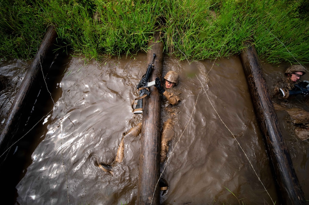 A person in a camouflage uniform with a rifle in one hand lies on their back in mud under a log and barbed wire.