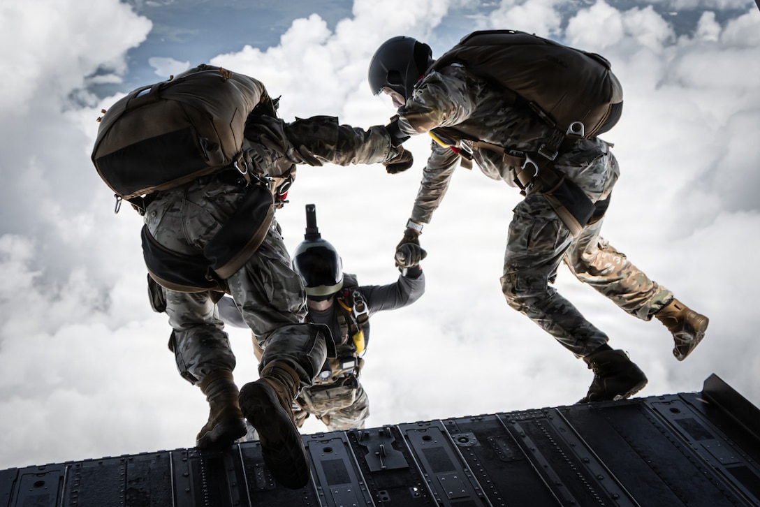 Three paratroopers link arms as two prepare to jump from an open aircraft to join a third in the sky.