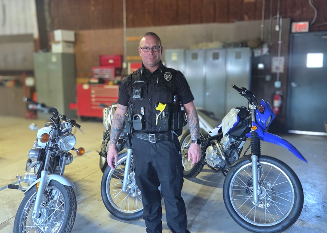 a male police officer in a blue police uniform stands inside a motor pool with three motorcycles behind him