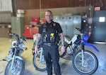 a male police officer in a blue police uniform stands inside a motor pool with three motorcycles behind him