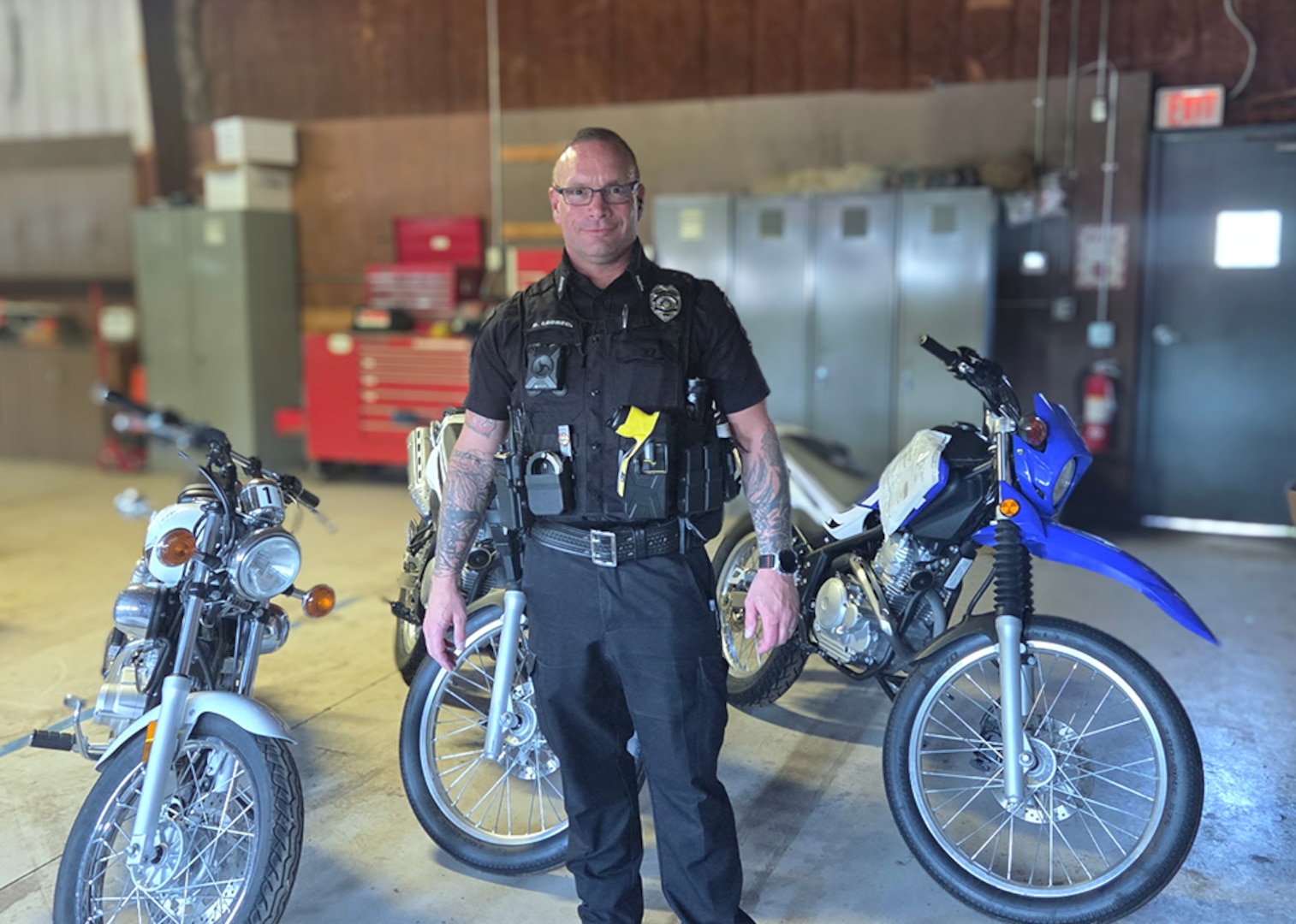 a male police officer in a blue police uniform stands inside a motor pool with three motorcycles behind him