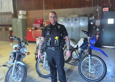 a male police officer in a blue police uniform stands inside a motor pool with three motorcycles behind him