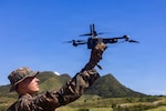 A man wearing a camouflage military uniform holds a military drone in the air as he stands in a grassy field with rolling hills in the background.
