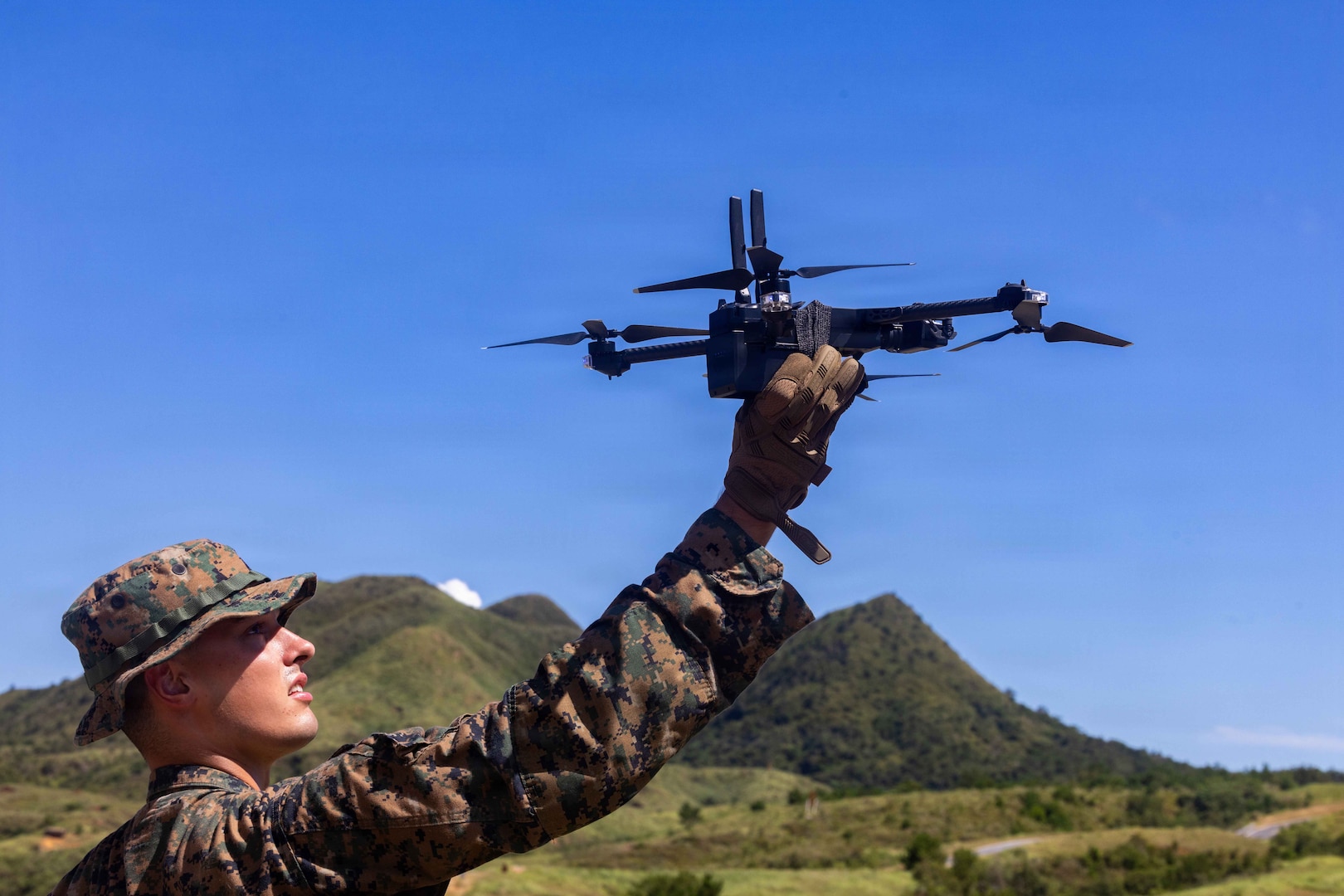 A man wearing a camouflage military uniform holds a military drone in the air as he stands in a grassy field with rolling hills in the background.