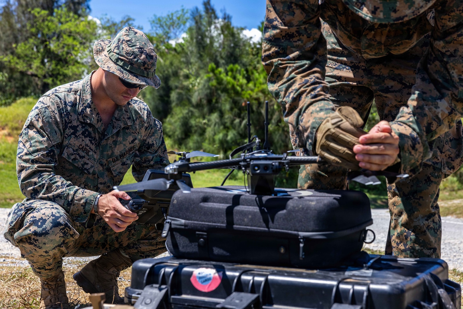 Two men wearing camouflage military uniforms crouch down to work on a miliary drone sitting on a black cargo box.