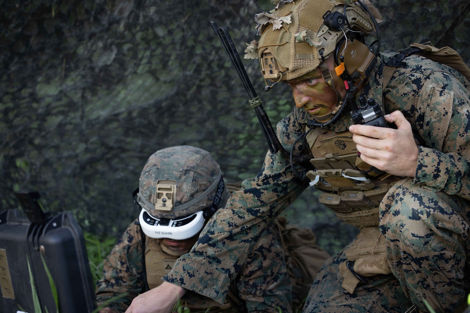 A man wearing a camouflage military uniform and face paint holds a satellite phone while looking at a computer screen. Another man in similar attire is wearing military goggles on his face.
