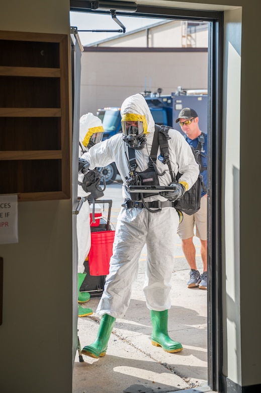 A man wearing a white hazmat suite and green boots stands just outside the entrance of a door. Another man in a blue shirt and khaki shorts stands behind him.