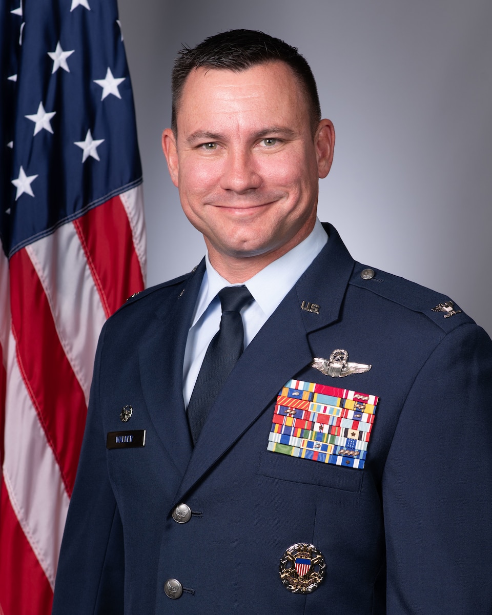An Airman wearing his dress blues stands in front of an American flag and a grey background