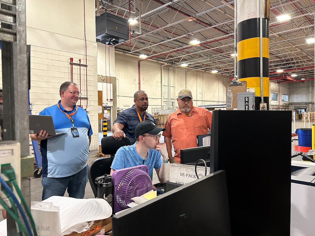 people stand around a computer monitor inside of a warehouse