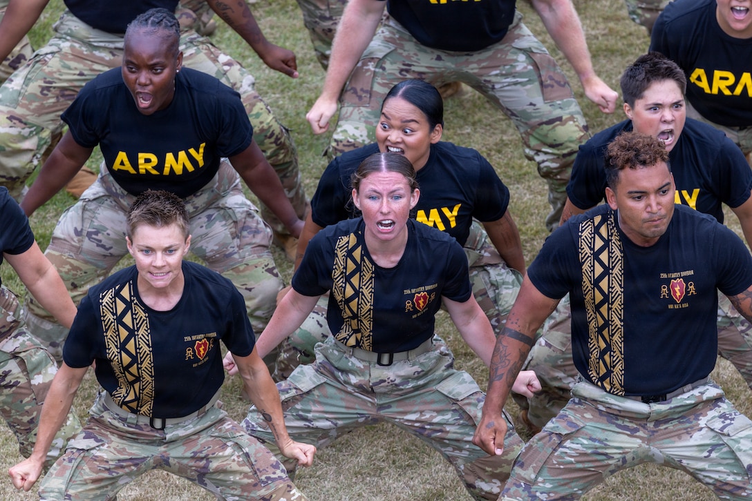 Soldiers in black T-shirts and camouflage pants perform a Hawaiian dance on a field as seen from slightly above.